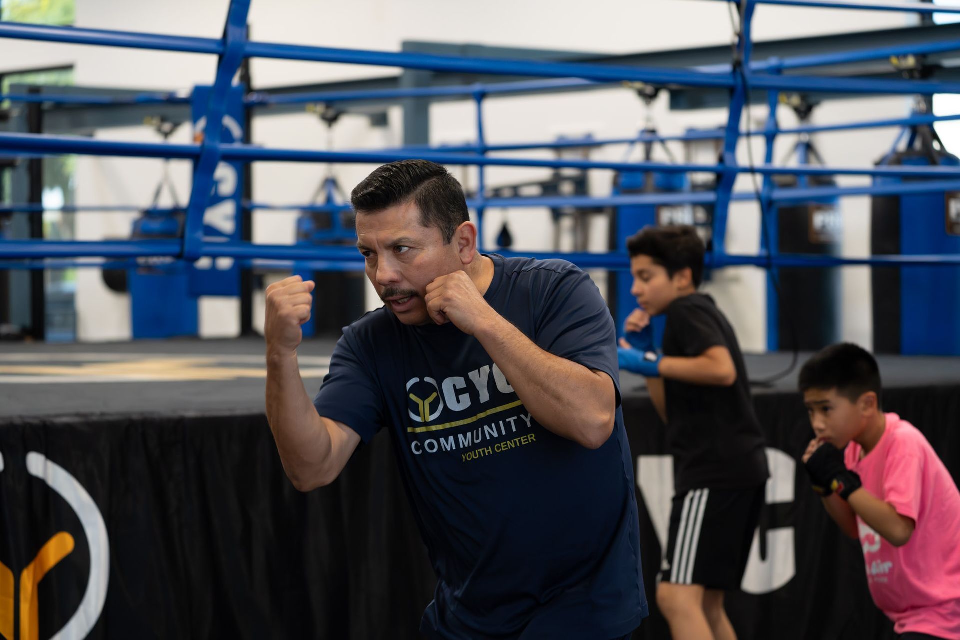 Boxer training in a gym, fists raised, with two youths practicing behind him in a boxing ring.