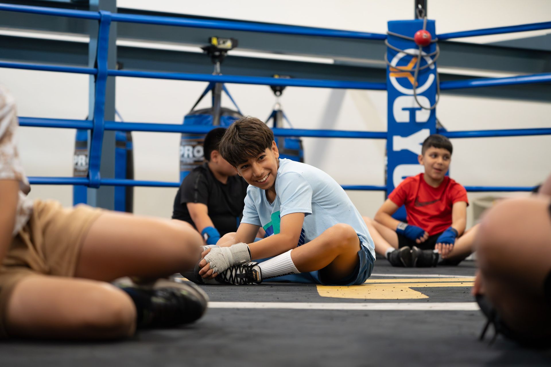 Boxing gym with two people stretching on the mat and others sitting nearby, blue ring ropes in the background