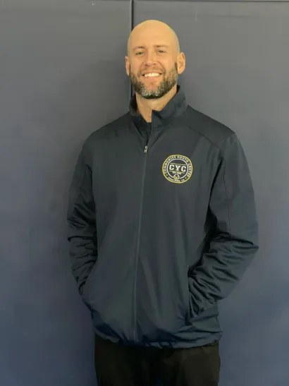 Smiling bald man in a black zip-up jacket with hands in pockets against a gray wall