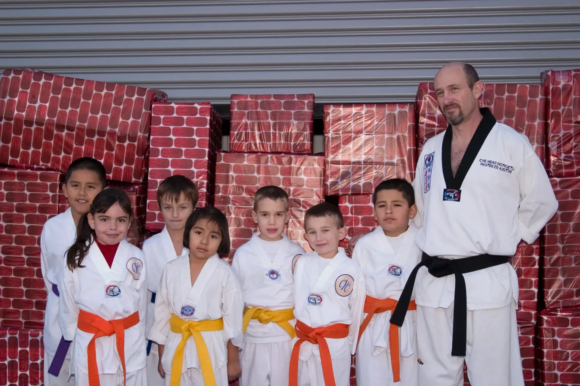 Martial arts instructor with six children in white uniforms and colored belts, standing in front of red gift boxes