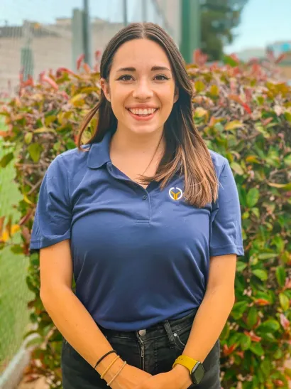 Smiling person in a blue polo shirt standing outdoors in front of colorful foliage.