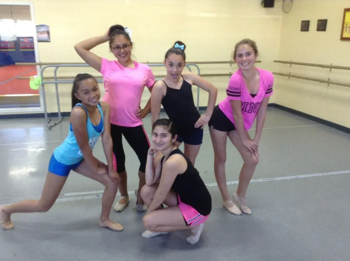 Five dancers posing in a studio, wearing bright practice outfits and smiling.