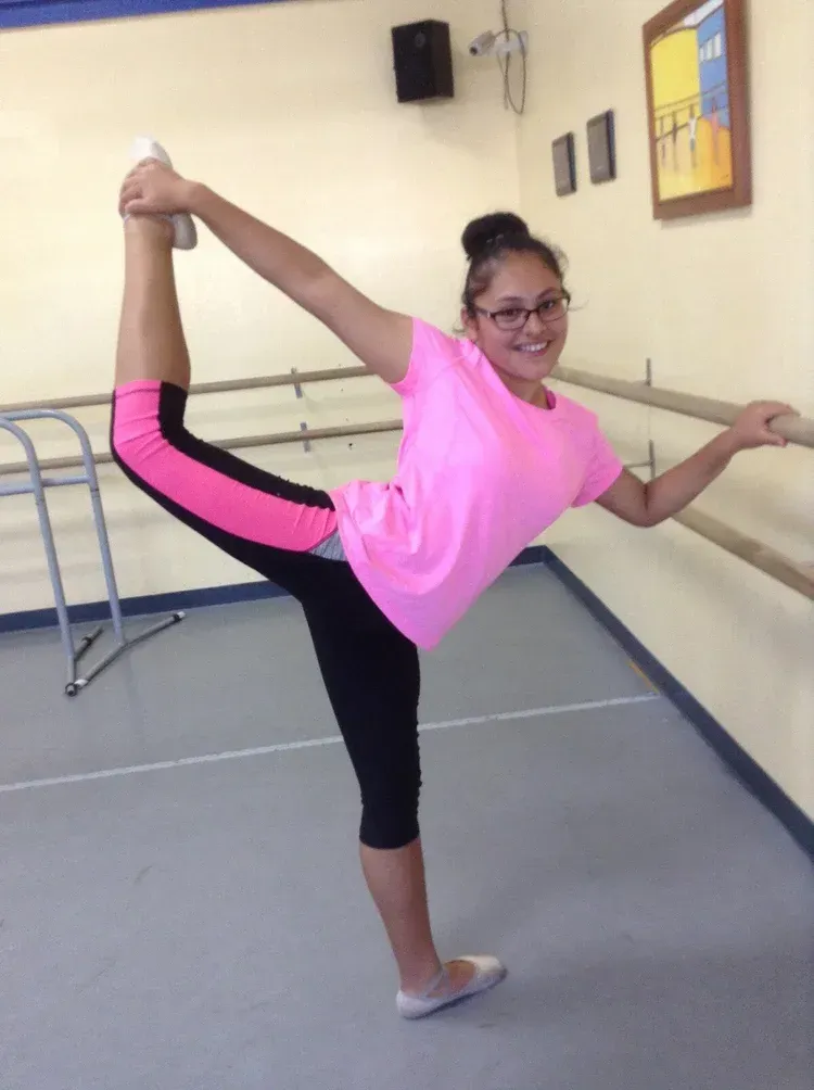 Girl in pink shirt and black leggings doing a ballet pose at a barre in a dance studio