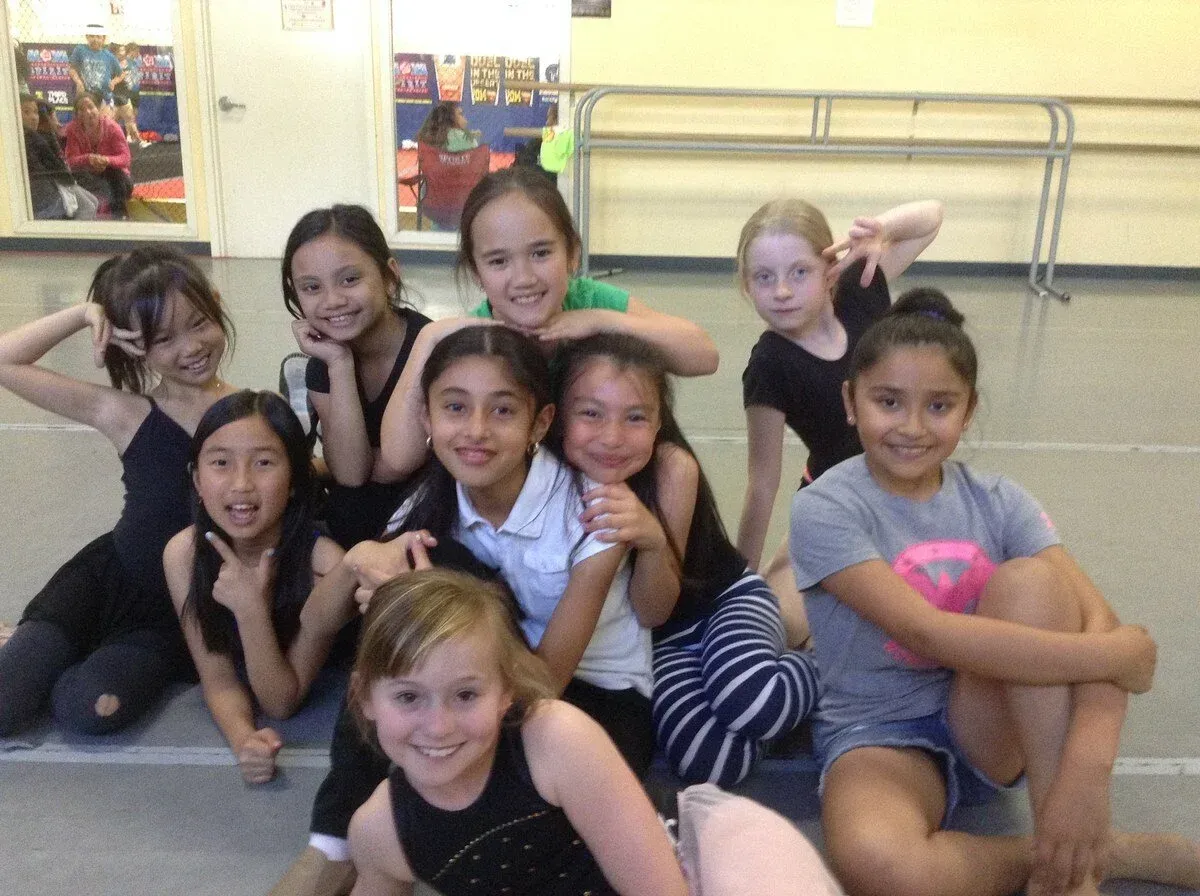 A group of children sitting on a studio floor, smiling and posing in dance costumes.