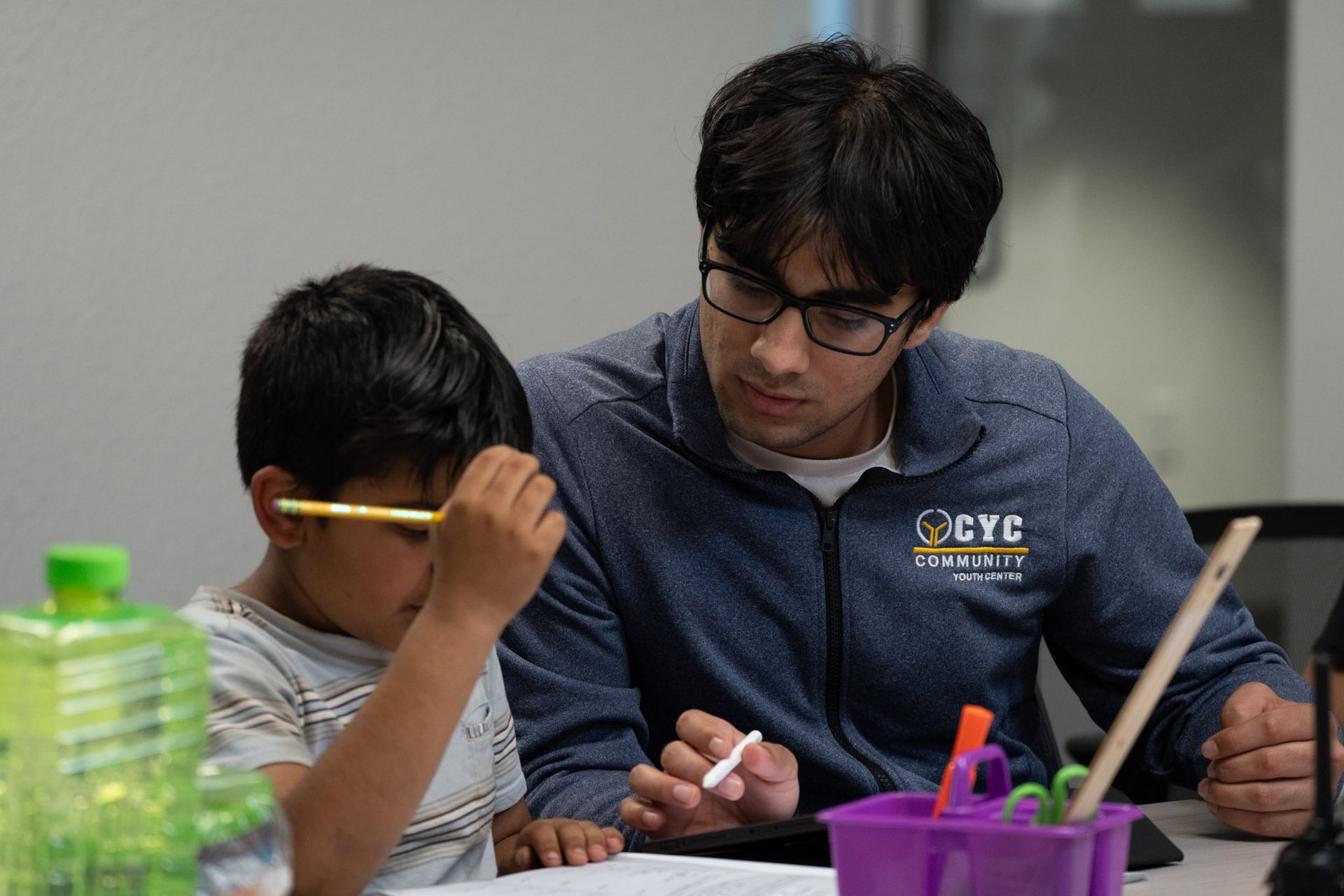 Two students working together at a table, one helping the other with a pencil in a classroom.