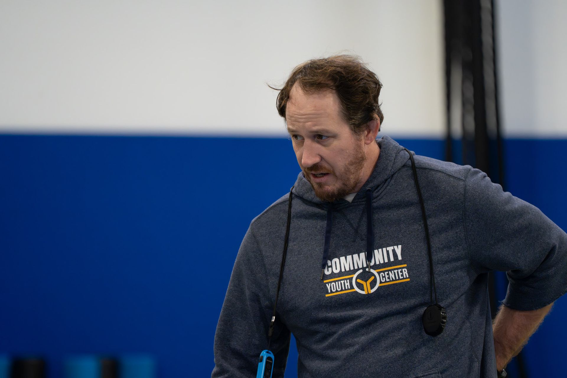 Man in a gray hoodie standing in a gym, looking down with a focused expression.