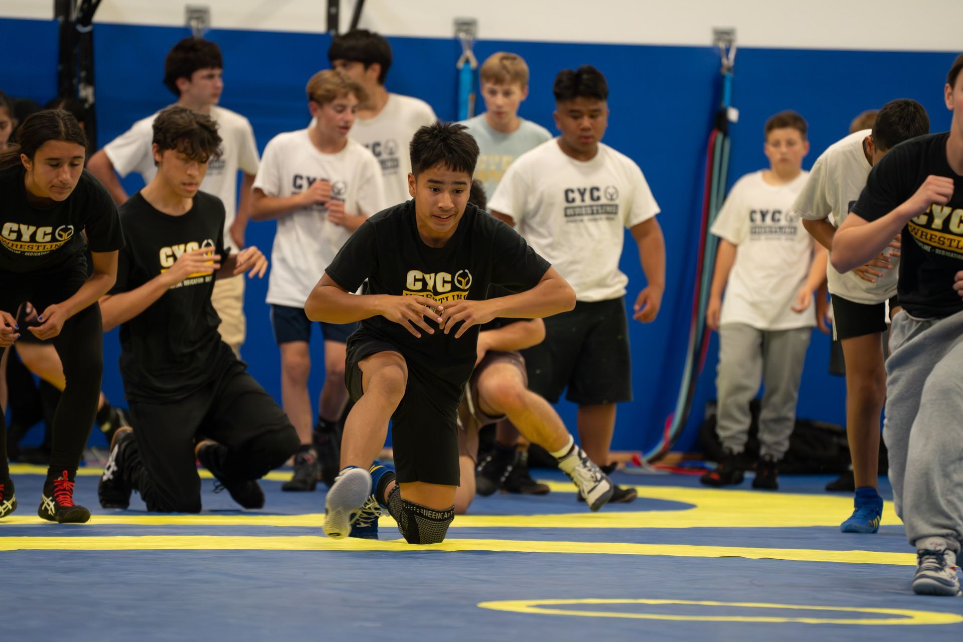 Wrestlers crouch on a mat during a match start, with teammates and spectators watching in the gym.