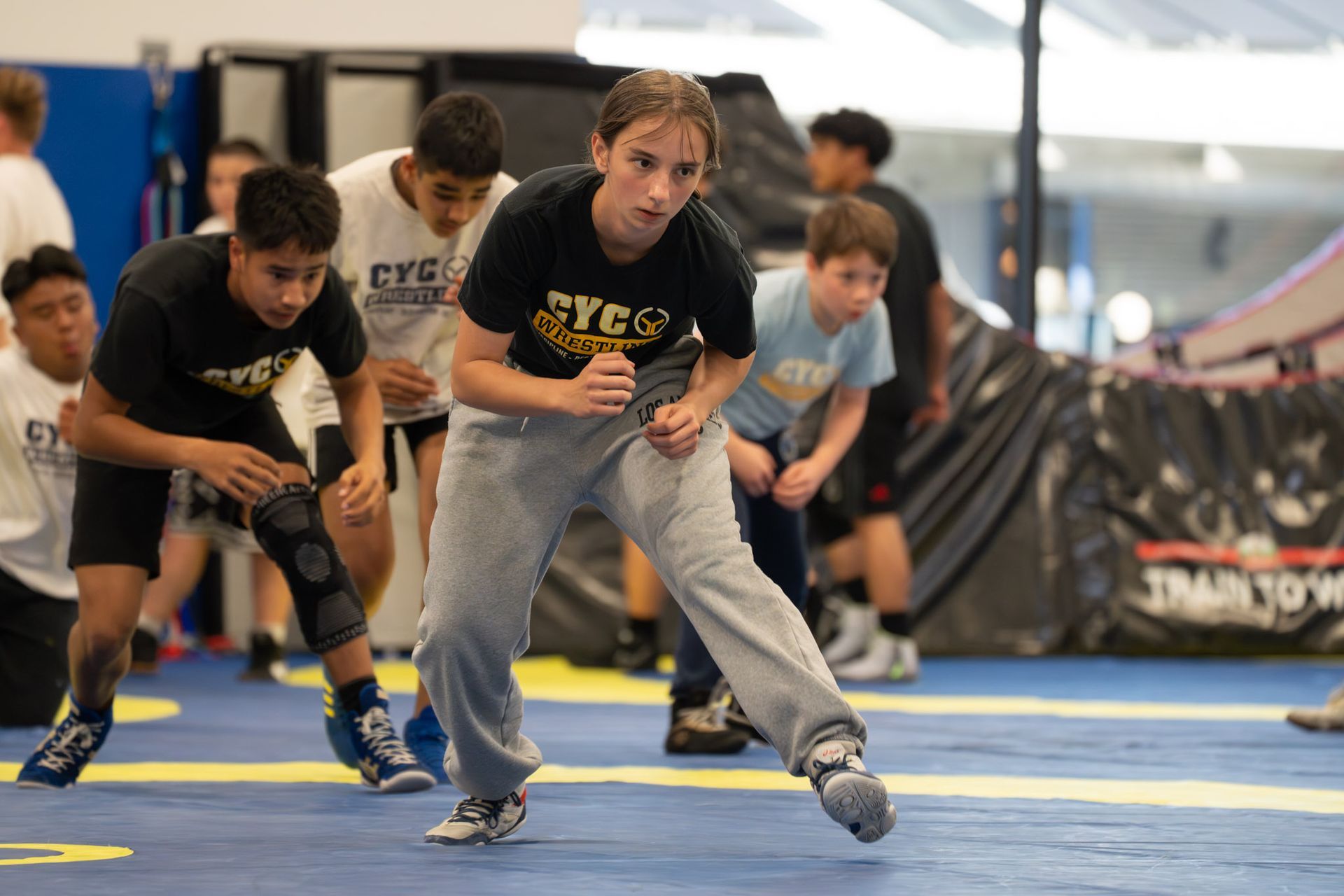 Children doing a wrestling drill on blue mats in a gym, leaning forward in ready stances