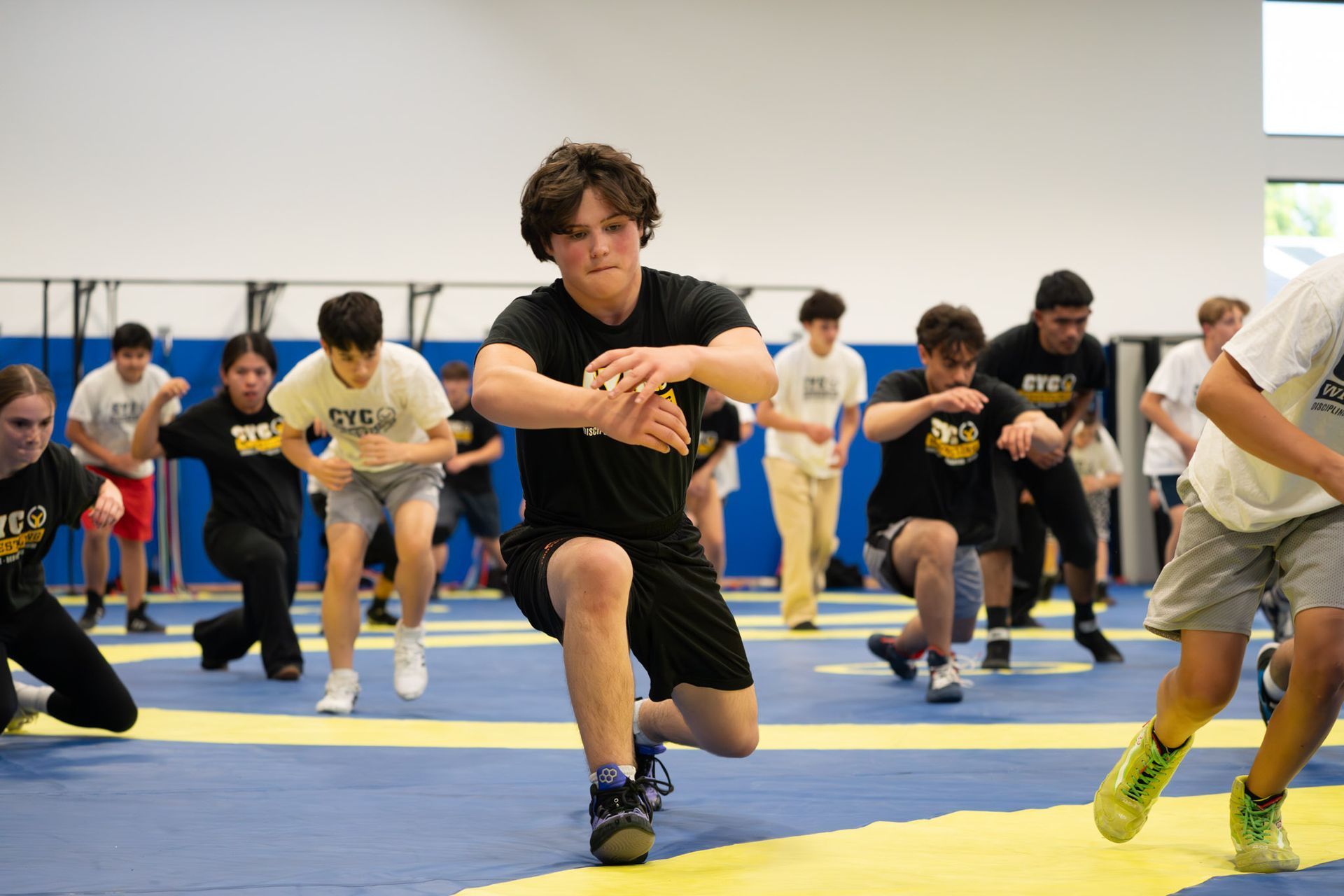 People exercising in a gym, lunging in unison on blue and yellow mats.