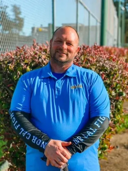 Man standing outdoors in a blue shirt, hands clasped, in front of bushes and a fence.