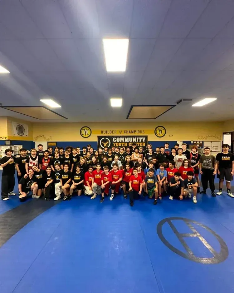 Large group posing in a martial arts gym with blue mats and a yellow wall banner.
