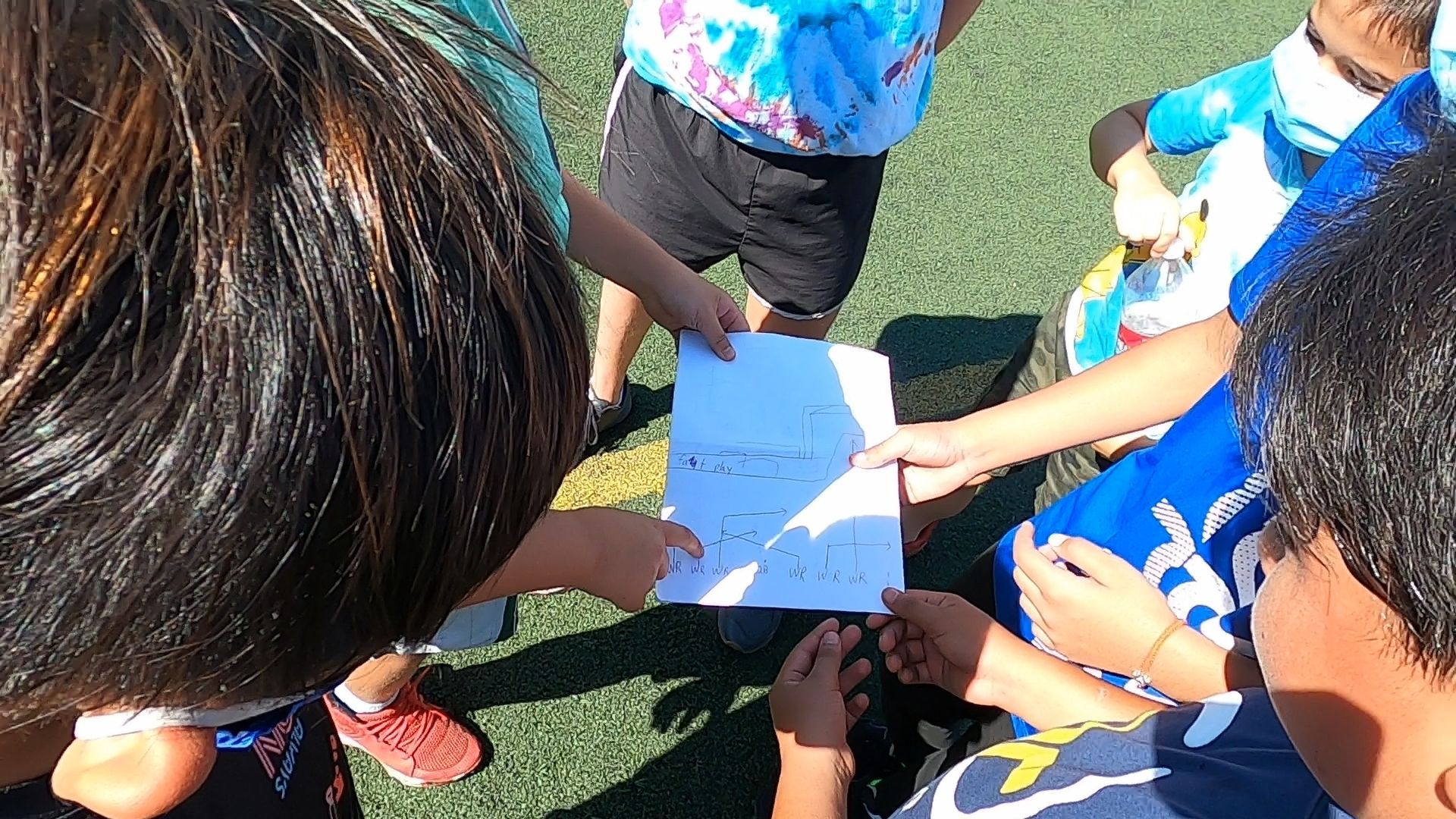 Children standing in a circle outdoors, reaching toward a paper on the grass.