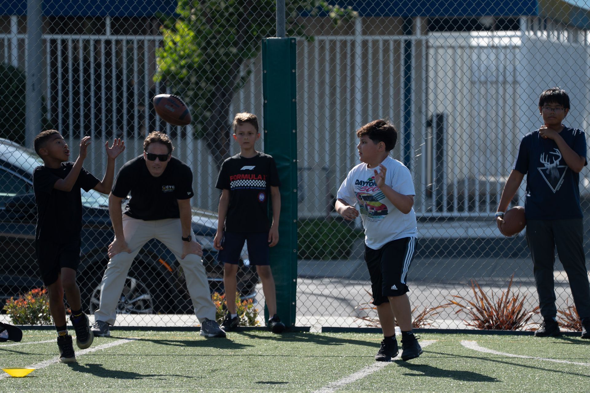 People playing football on an outdoor field near a chain-link fence in bright sunlight