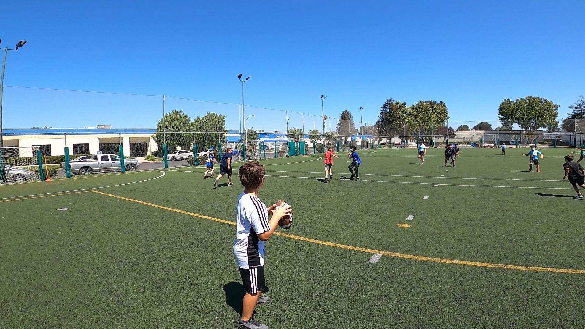 Youth soccer game on a green field, player holding a ball in the foreground under a clear blue sky.