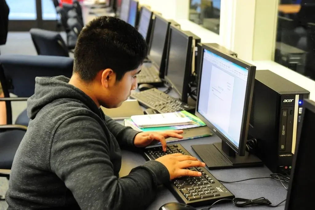 Person typing at a desktop computer in a row of monitors at a computer lab.
