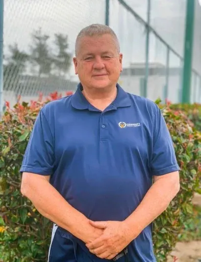Man in a blue polo shirt standing outdoors in front of shrubs and a fence.