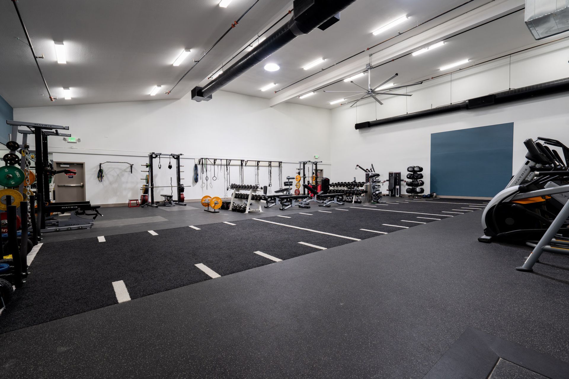 Empty indoor gym with black rubber flooring, white walls, and exercise equipment along the back wall