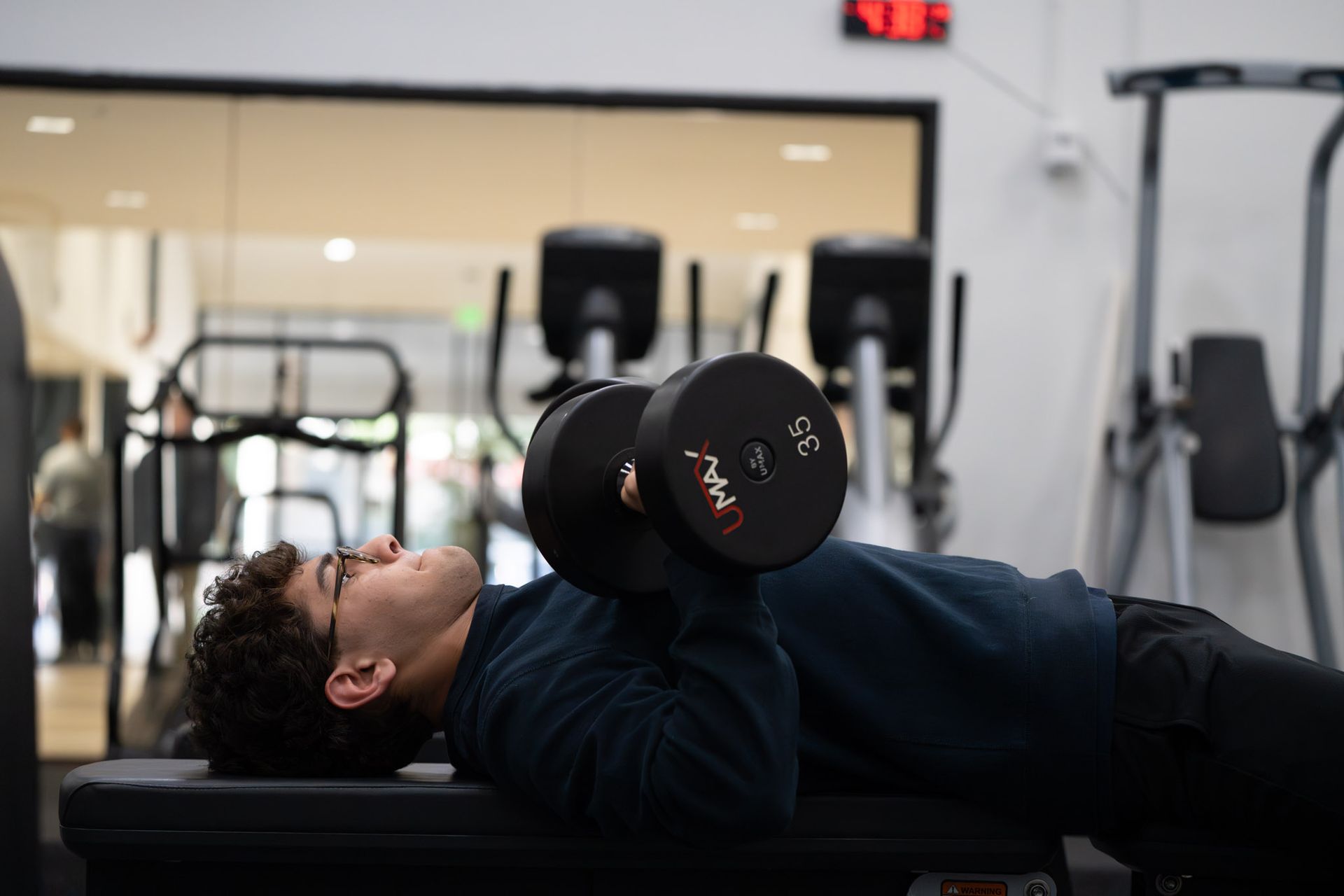 Person lying on a bench press, holding dumbbells in a gym.