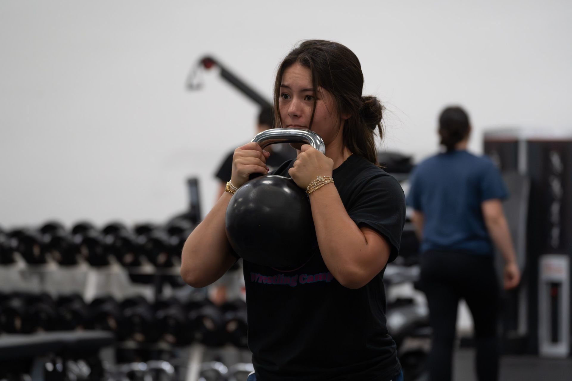 Person in a gym holding a kettlebell at chest height, with dumbbells and equipment in the background.