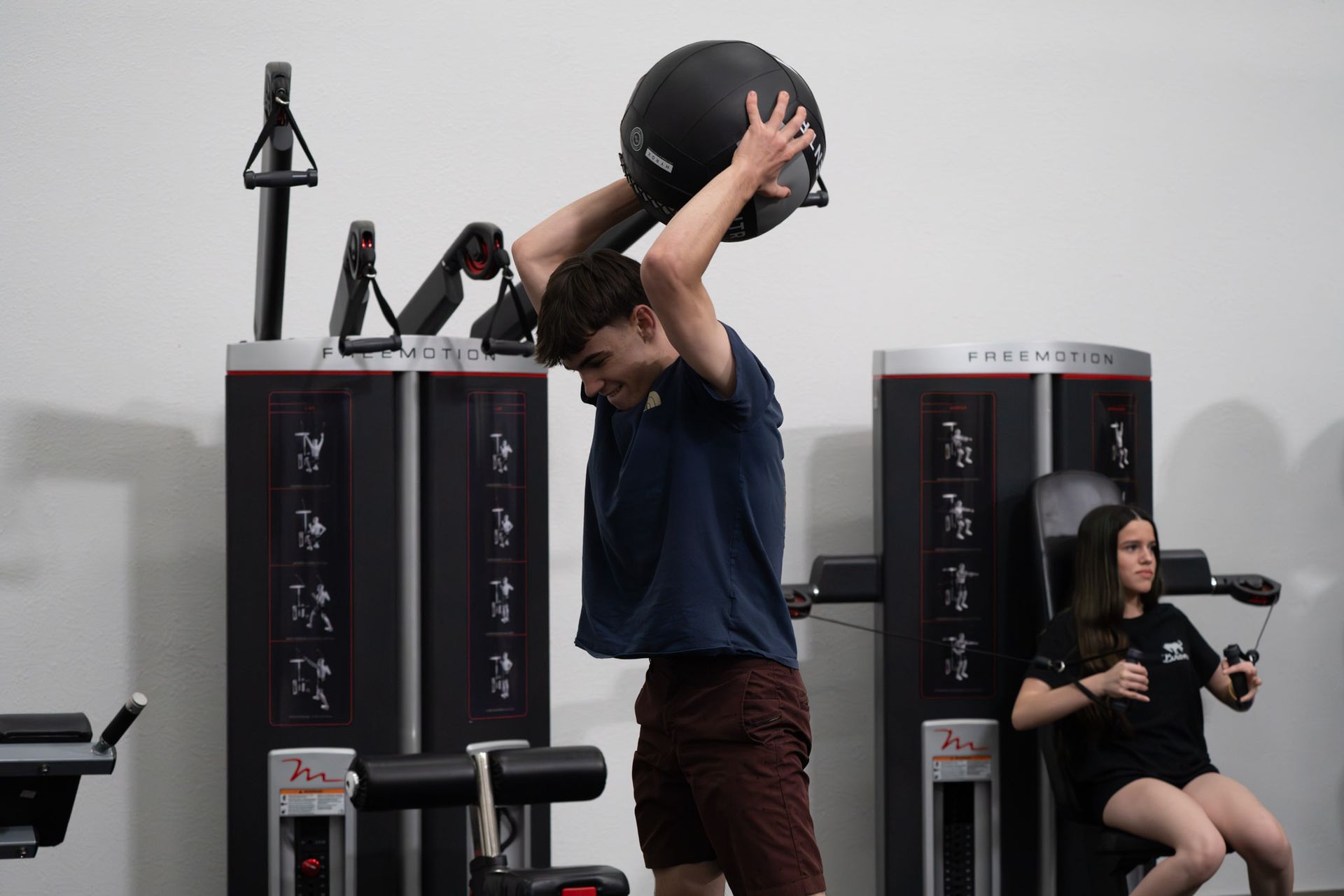 Man lifting a medicine ball overhead in a gym, with exercise machines in the background.