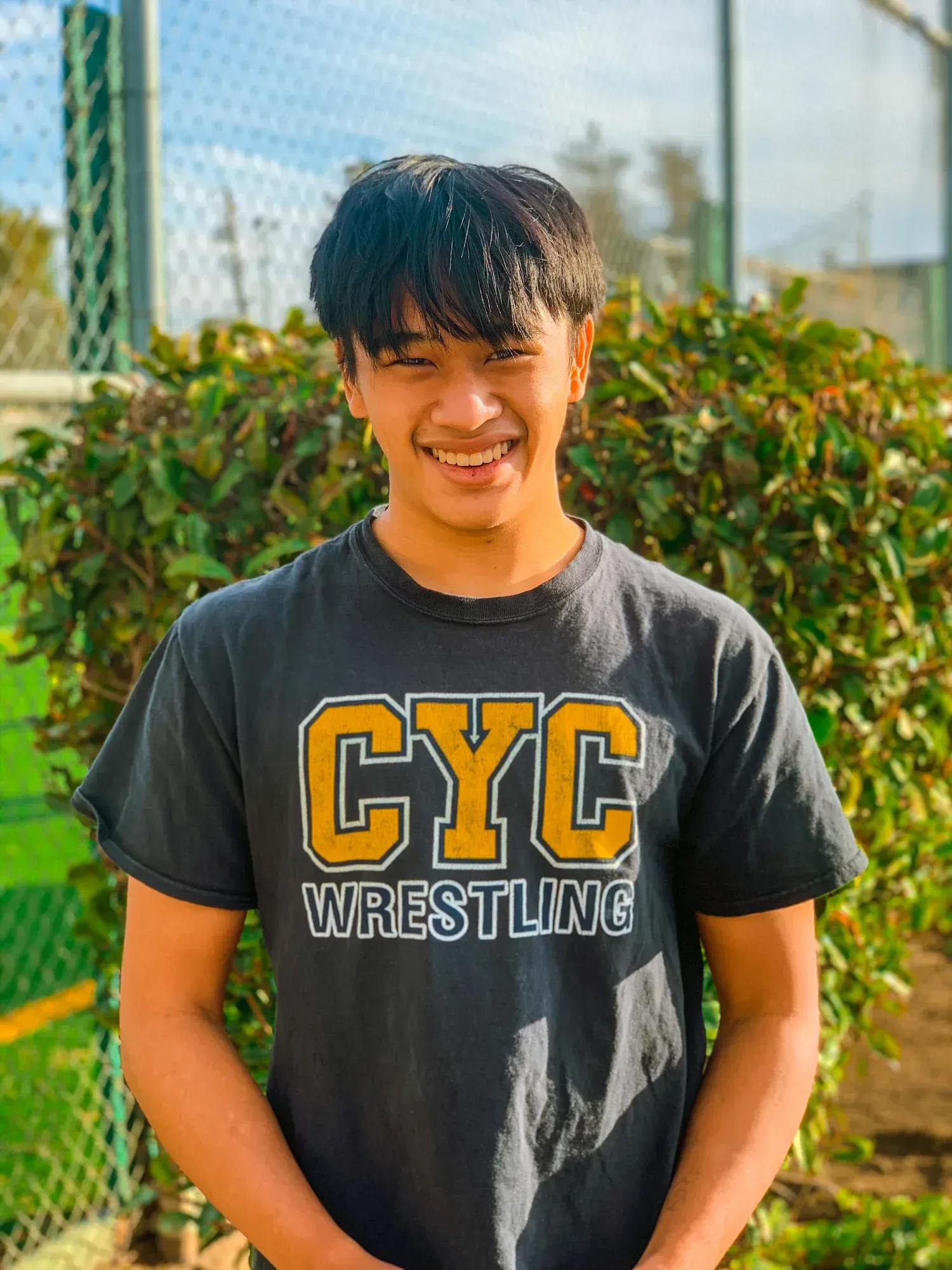 Smiling person in a black CYC Wrestling T-shirt standing outdoors near a chain-link fence.