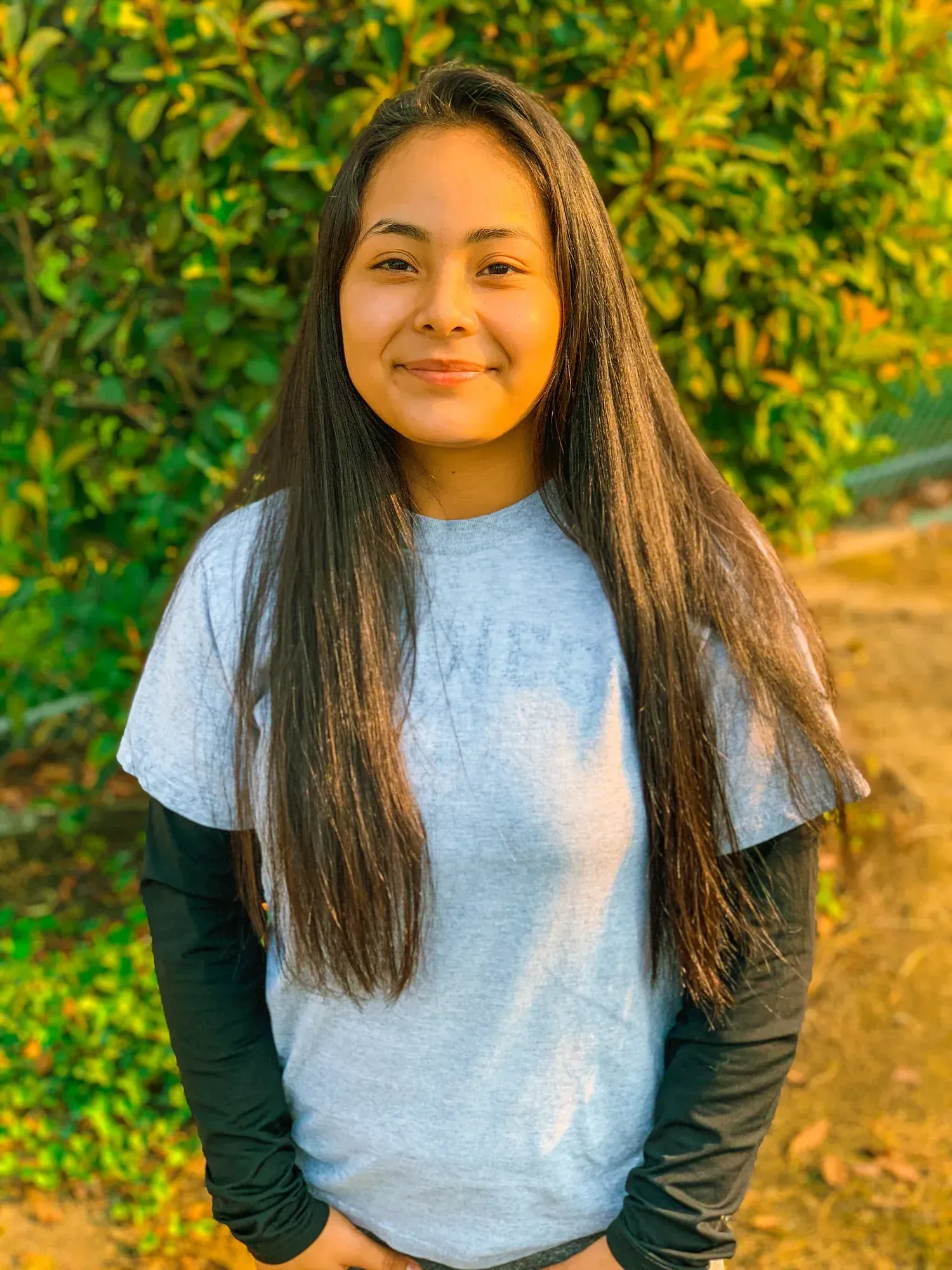 Smiling person with long dark hair standing outdoors in front of green foliage, wearing a gray T-shirt and black sleeves