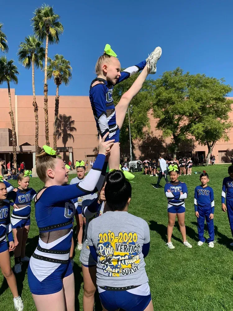 Cheerleaders in blue uniforms perform an outdoor stunt on a grassy field with palm trees and spectators nearby.
