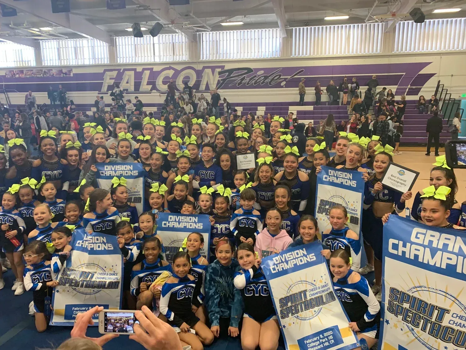 Large group of cheerleaders in blue uniforms holding “Game Champions” signs in a gym with a “Falcons” banner