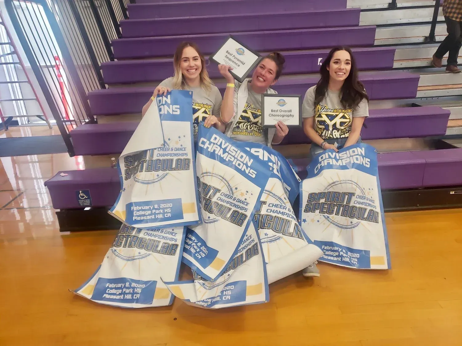 Three people on purple steps hold blue-and-white bags and plaques, smiling behind a large stack of awards.
