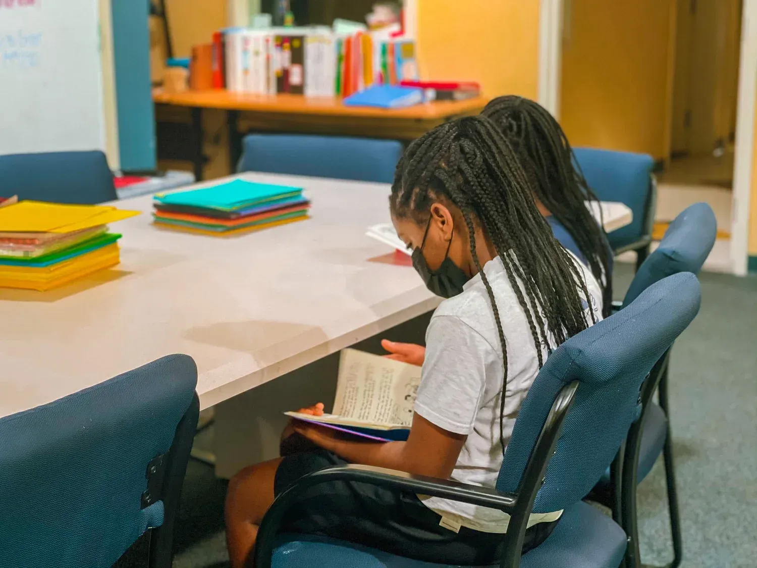 Student writing at a conference table in a classroom, with colorful notebooks stacked nearby.