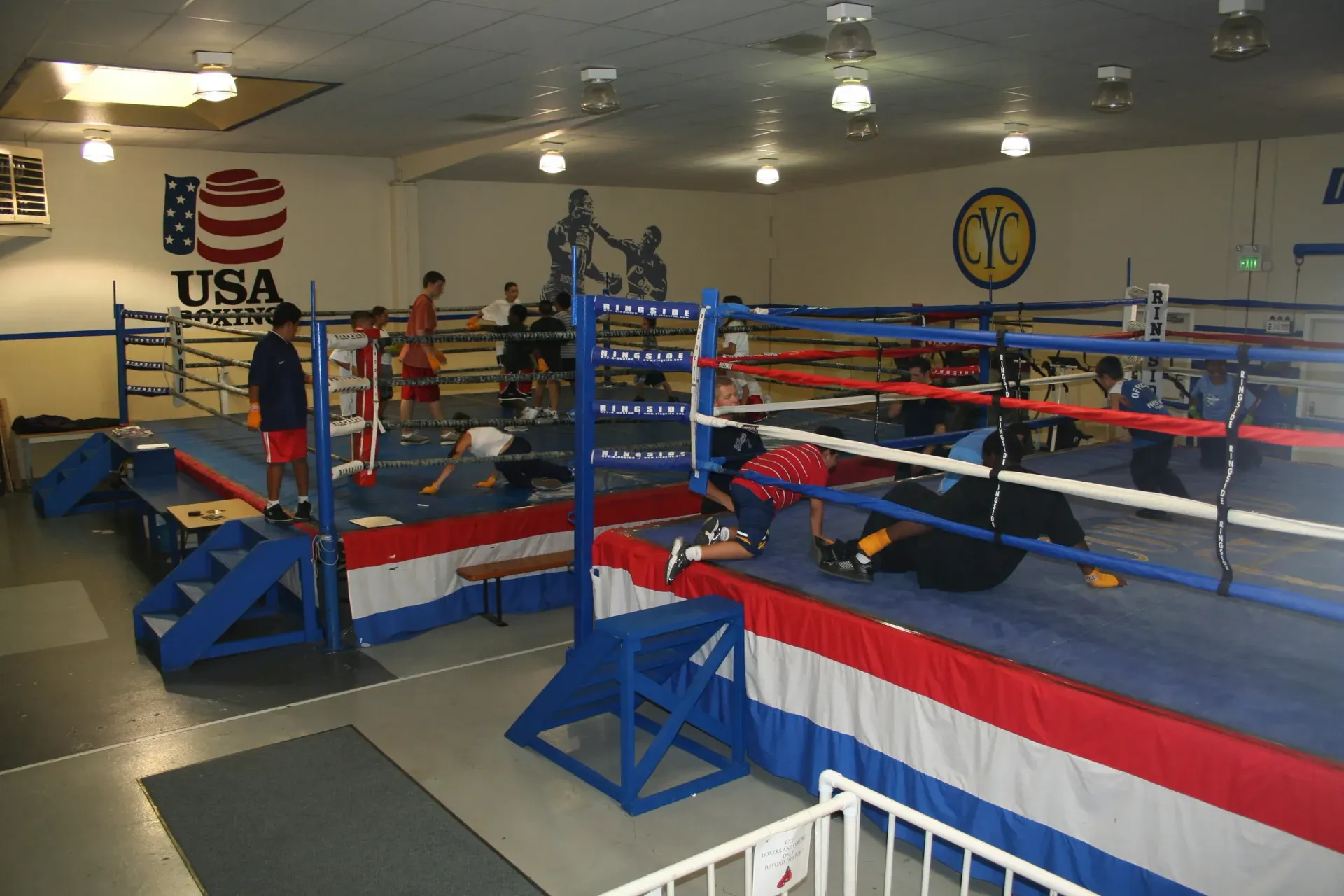 Boxing ring with red, white, and blue ropes in a gym, with training equipment and USA boxing sign on the wall