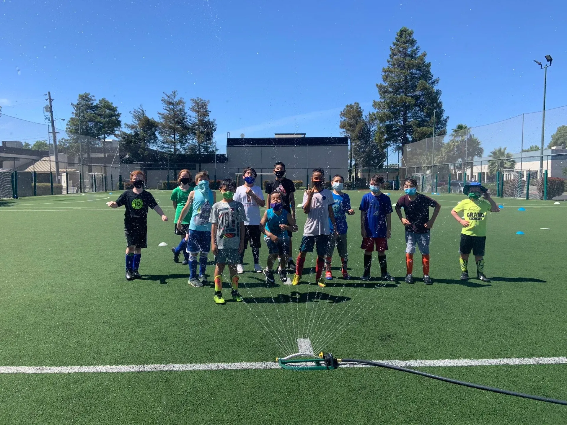 Youth soccer team lined up on a turf field, posing in bright sunlight with goals and trees behind them.