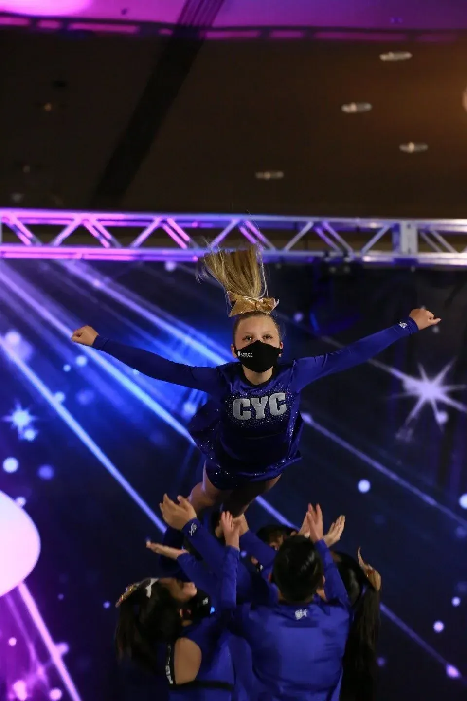 Cheerleader in blue uniform is tossed high by teammates on a lit stage under purple lights