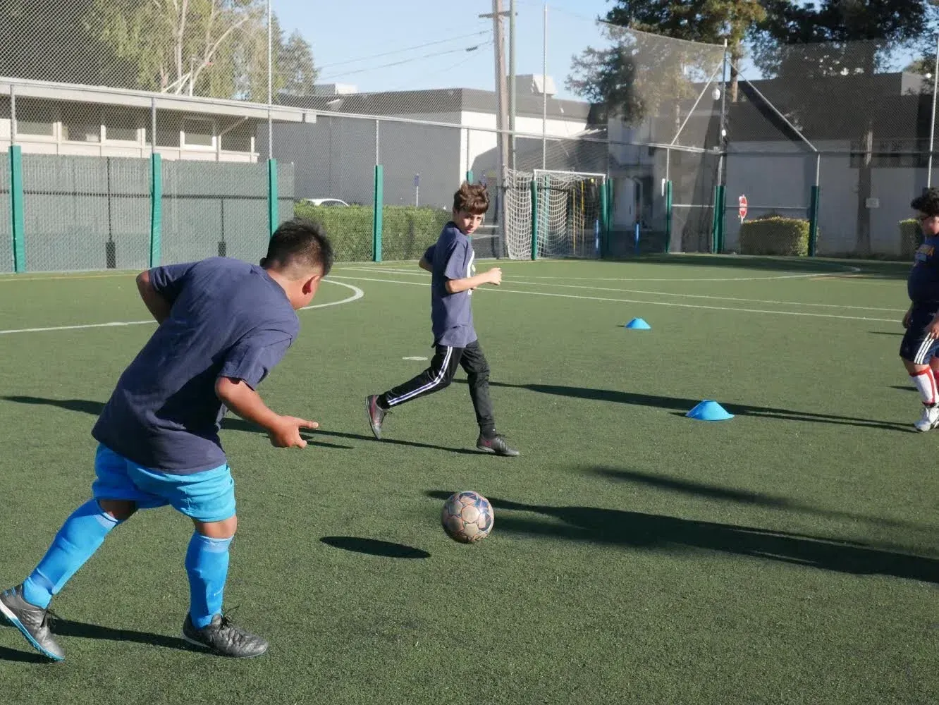Children playing soccer on an outdoor field with blue cones and a ball near the goal.