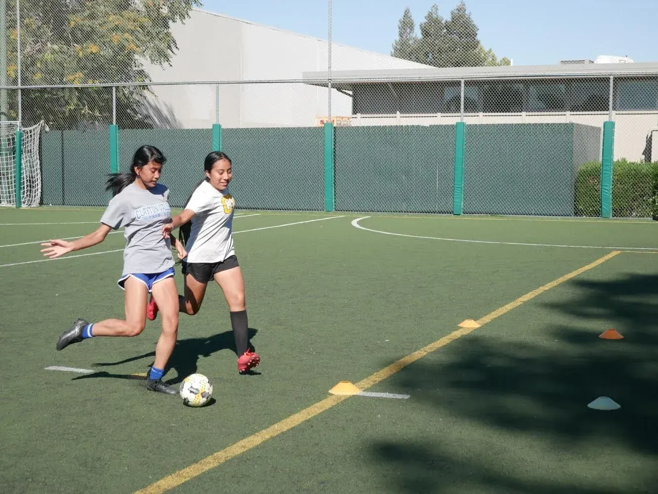 Two people playing soccer on an outdoor turf field, one kicking the ball near the goal.