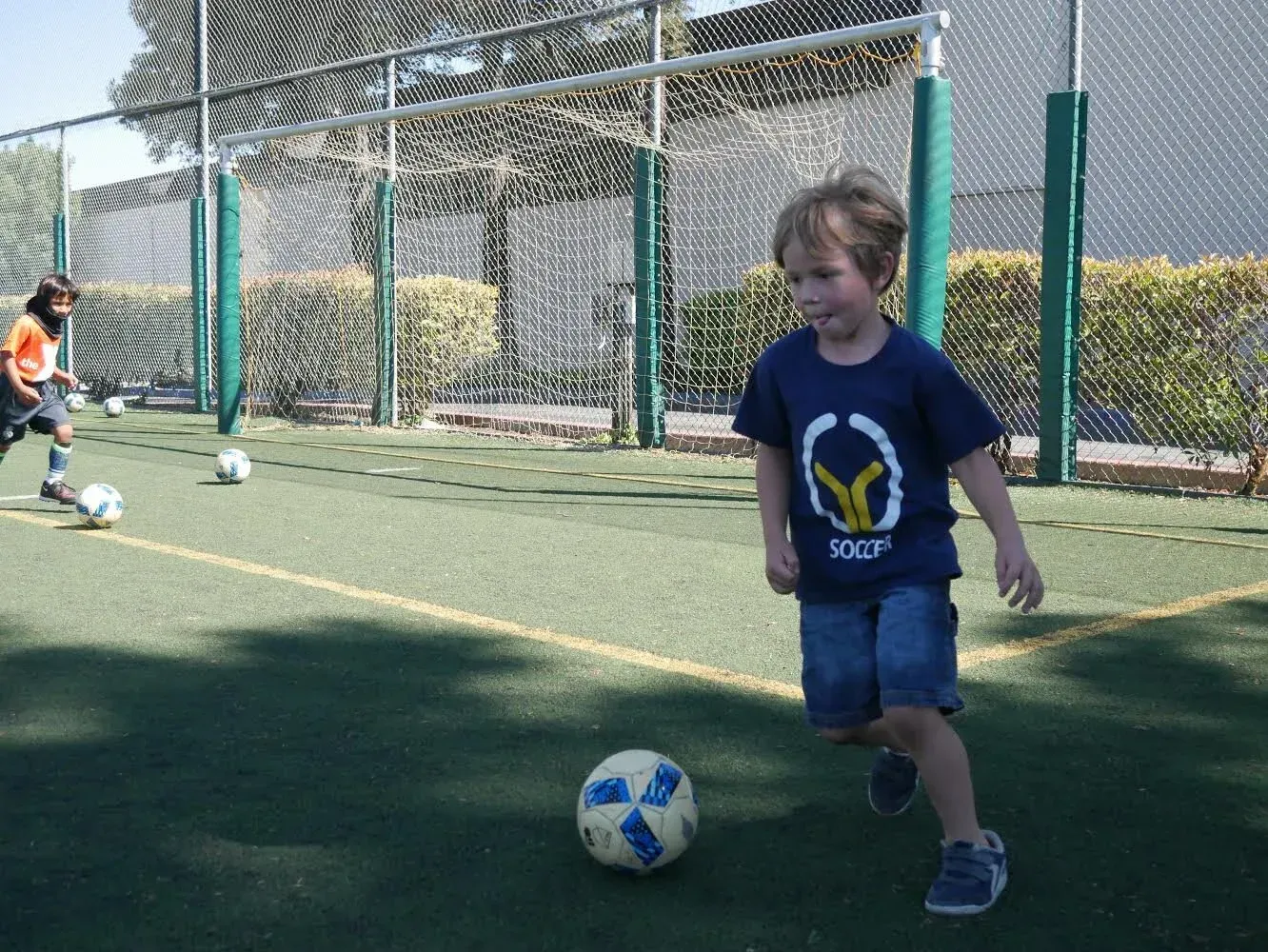 Child dribbling a soccer ball on an outdoor field, with another child and goal in the background
