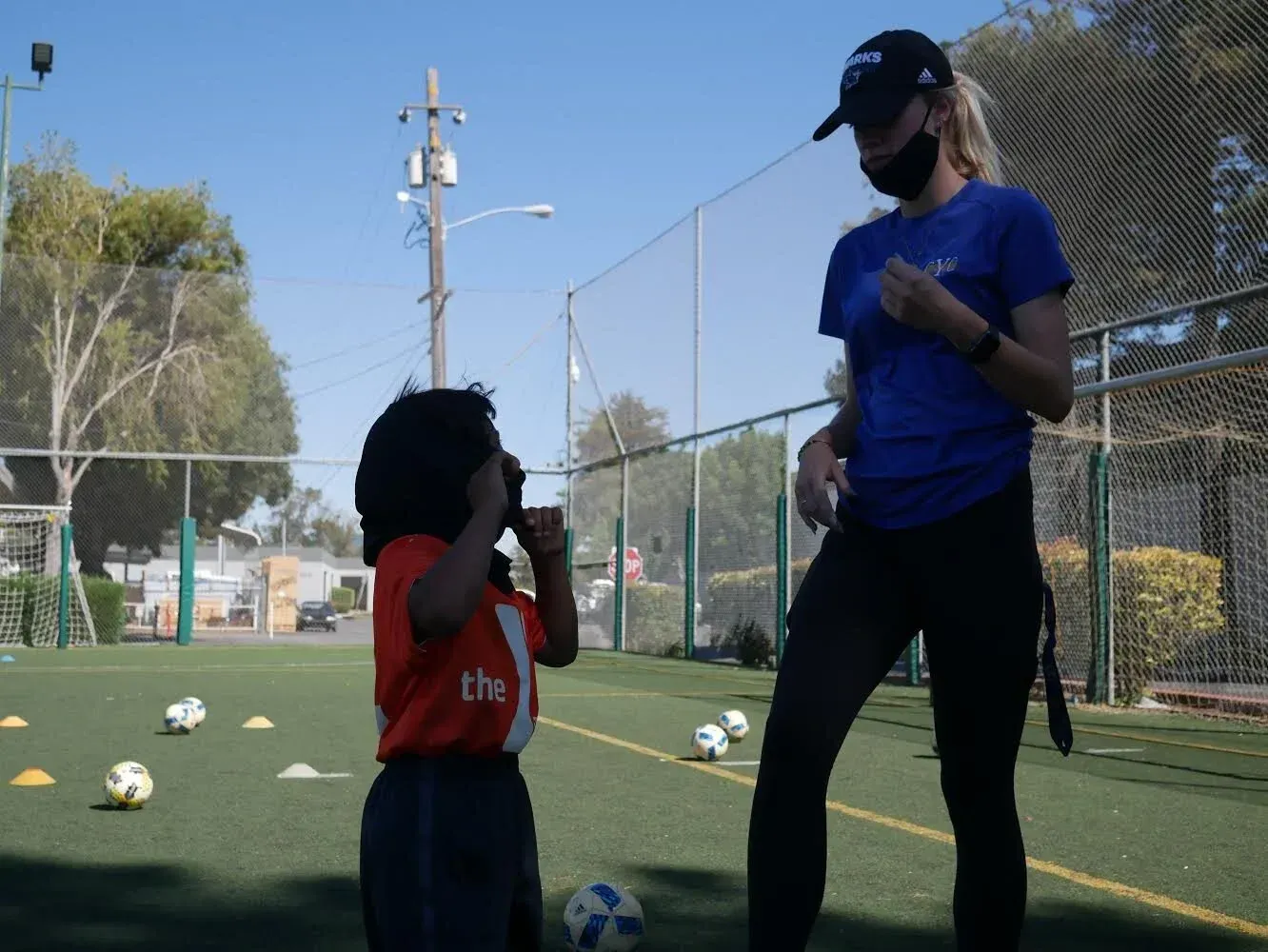 Coach and child standing on a soccer field, talking near scattered balls and a goal net.
