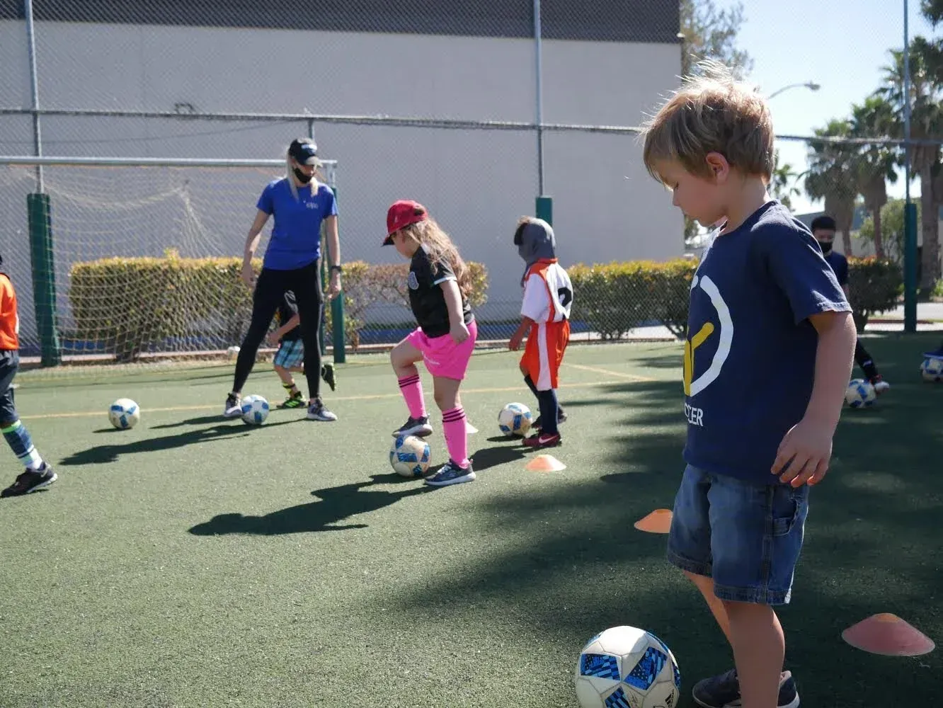 Children practicing soccer cones on an outdoor turf field with a coach nearby