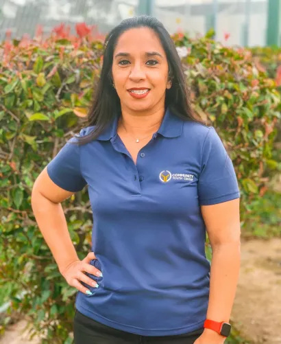 Woman in a blue polo shirt standing outdoors, smiling with hands on hips in front of greenery.