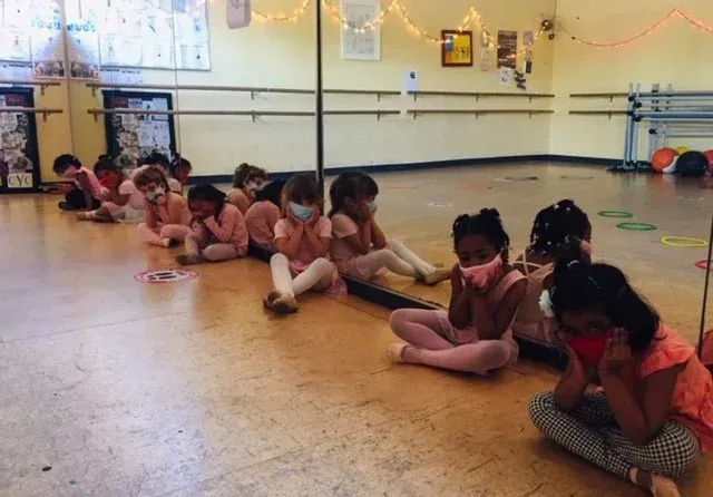 Children seated on the floor in a dance studio, stretching during a group class.