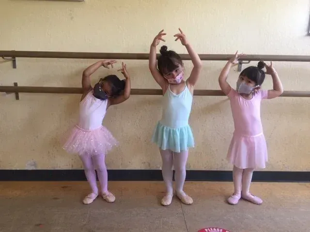 Three children in pastel dance outfits pose with arms raised at a ballet studio barre.