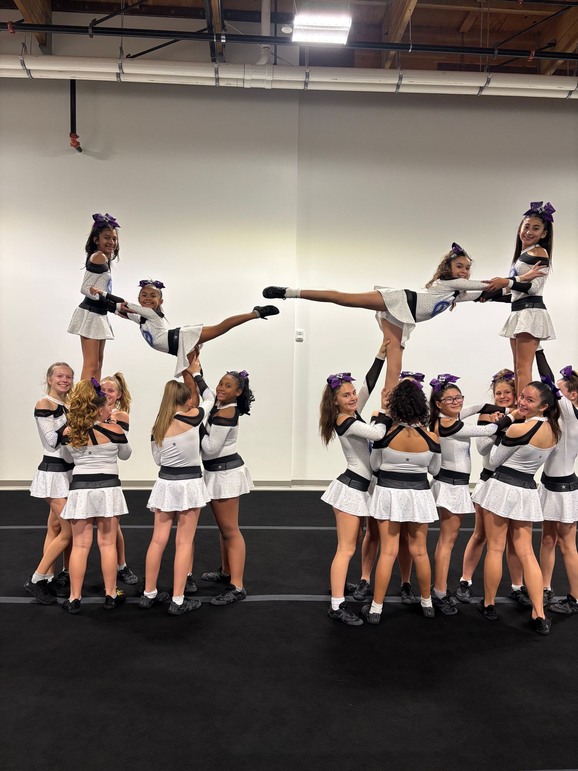 Cheerleaders in black and white uniforms performing a stunt pyramid indoors, with two flyers extended horizontally.
