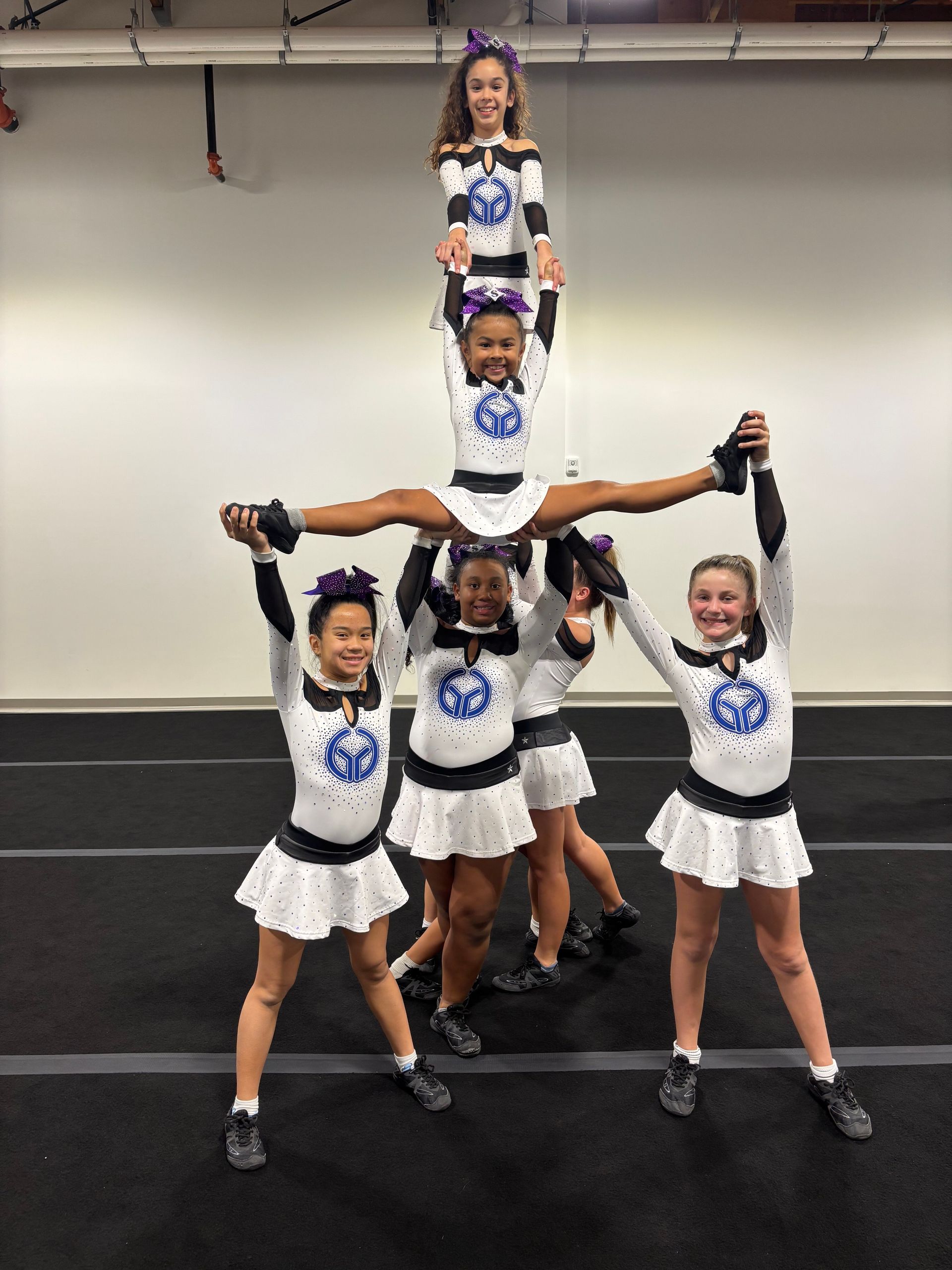 Six cheerleaders in white uniforms pose in a human pyramid in a gym.