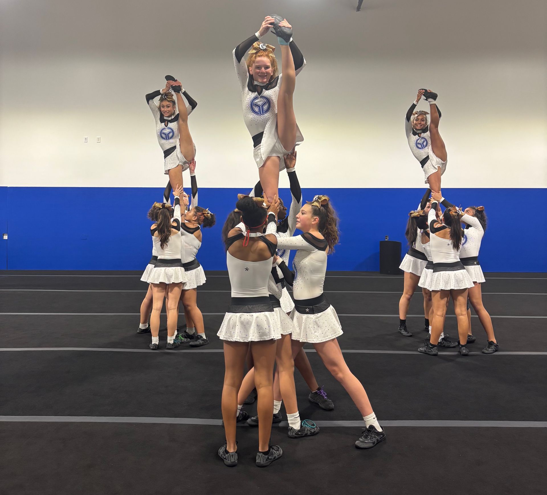 Cheerleaders performing stunts in a gym, with one flyer held overhead in the center and two groups on the sides.