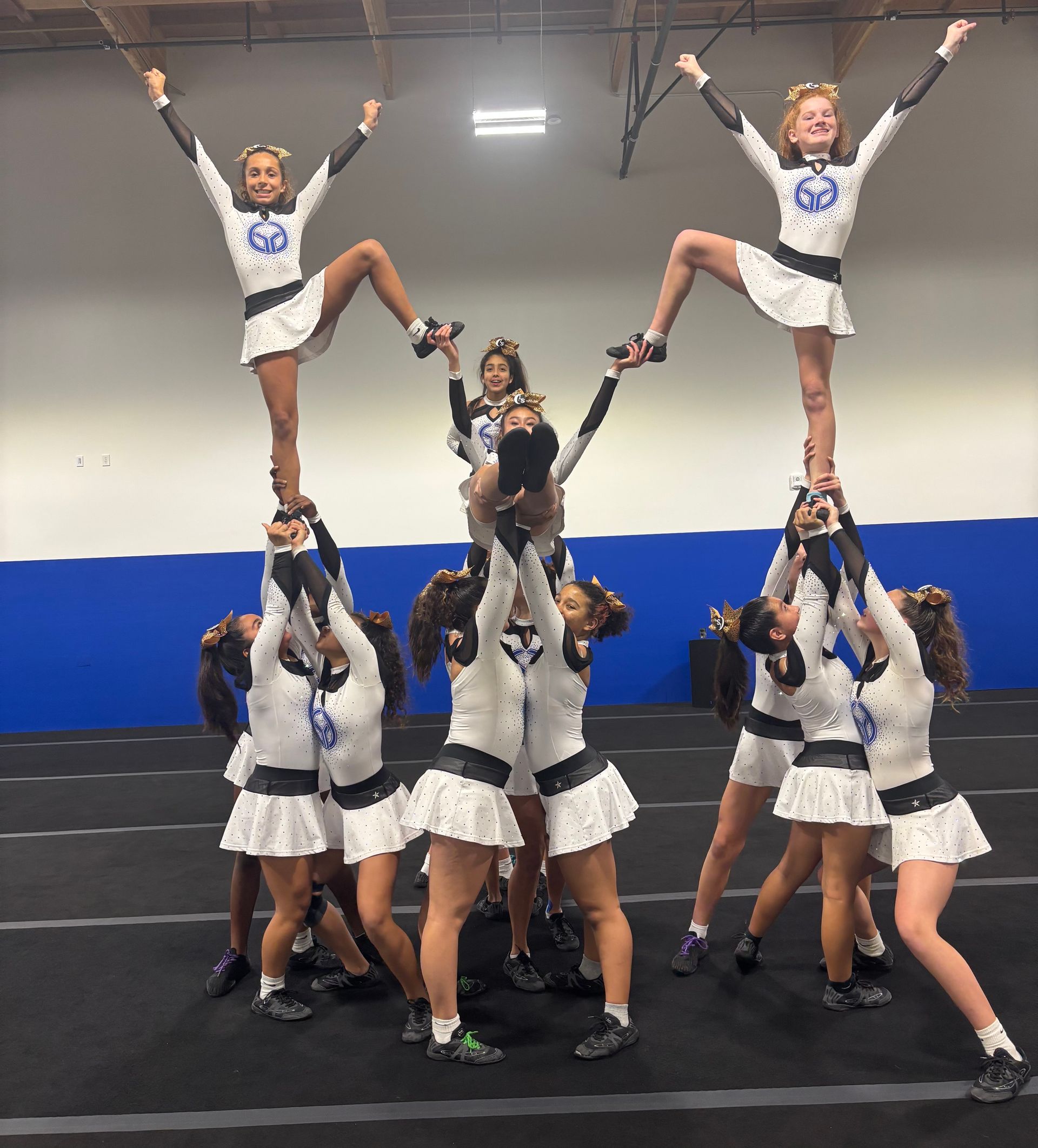 Cheerleaders in white and blue uniforms performing a pyramid stunt indoors on a mat