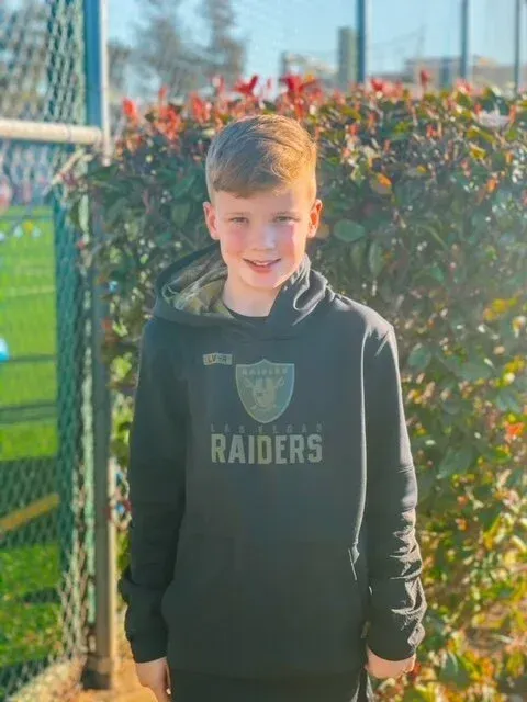 Boy standing outdoors by a fence, wearing a black Raiders hoodie, in bright sunlight