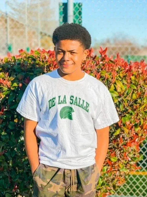 Person in a white De La Salle T-shirt standing outdoors in front of colorful bushes.