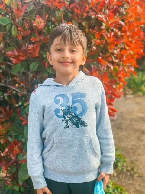 Child in a light blue hoodie with a dinosaur print, standing in front of red autumn leaves.