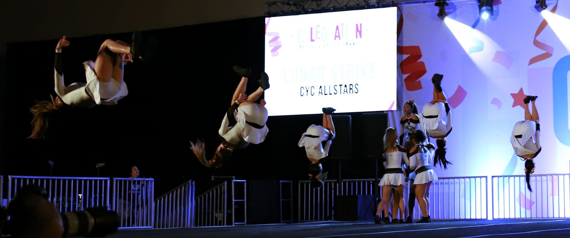 Cheerleaders performing jumps on a lit stage with a projected sign in the background