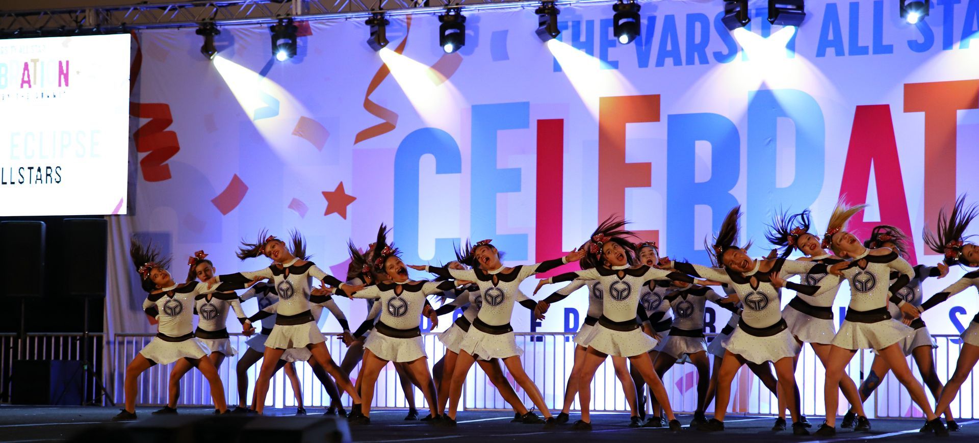 Dancers in white costumes perform on a colorful stage with large “CELEBRATE” lettering and confetti backdrop
